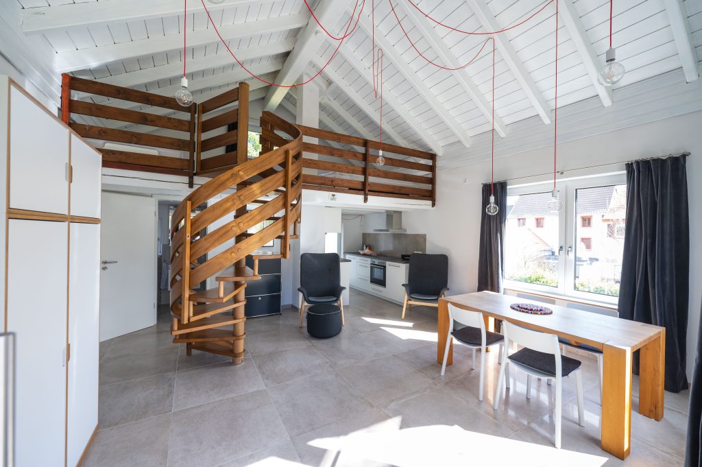 Kleines Tiny Ferienhäuschen mit grauen Fensterrahmen und dunklem Ziegeldach inmitten einer gepflegten Grünfläche. Vor dem Haus steht eine gemütliche Steinterrasse mit Gartenmöbeln, umgeben von Büschen, Bäumen und einem blauen Himmel mit wenigen Wolken.